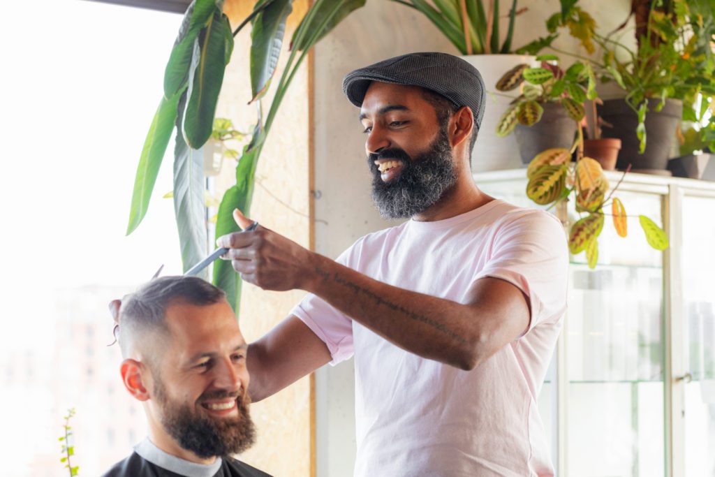 Ein lächelnder Barbier mit dunklem Bart und einer grauen Schiebermütze schneidet einem lächelnden Kunden in einem hellen, mit grünen Zimmerpflanzen dekorierten Salon die Haare mit Schere und Kamm.