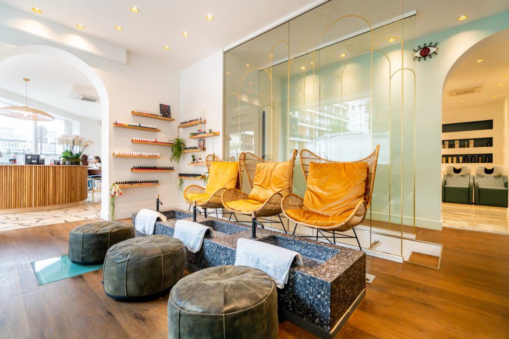 Elegantes, helles Interieur eines Schönheitssalons mit einem speziellen Pediküre-Bereich, der drei senfgelbe Korbsessel, Steinbecken mit weißen Handtüchern und passende graue Hocker auf einem Holzboden umfasst.