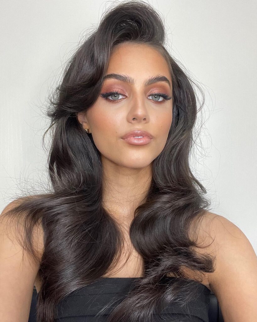 A woman with long, dark wavy hair featuring elegant side-swept bangs angled to one side to soften facial contours.