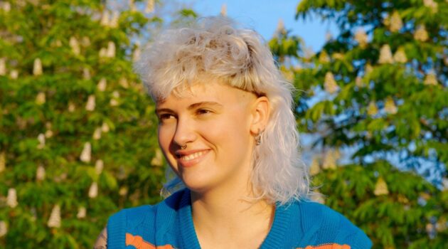Eine junge Frau mit lockigem, wei&szlig;blondem Vokuhila-Haarschnitt, die an einem sonnigen Tag drau&szlig;en sitzt.