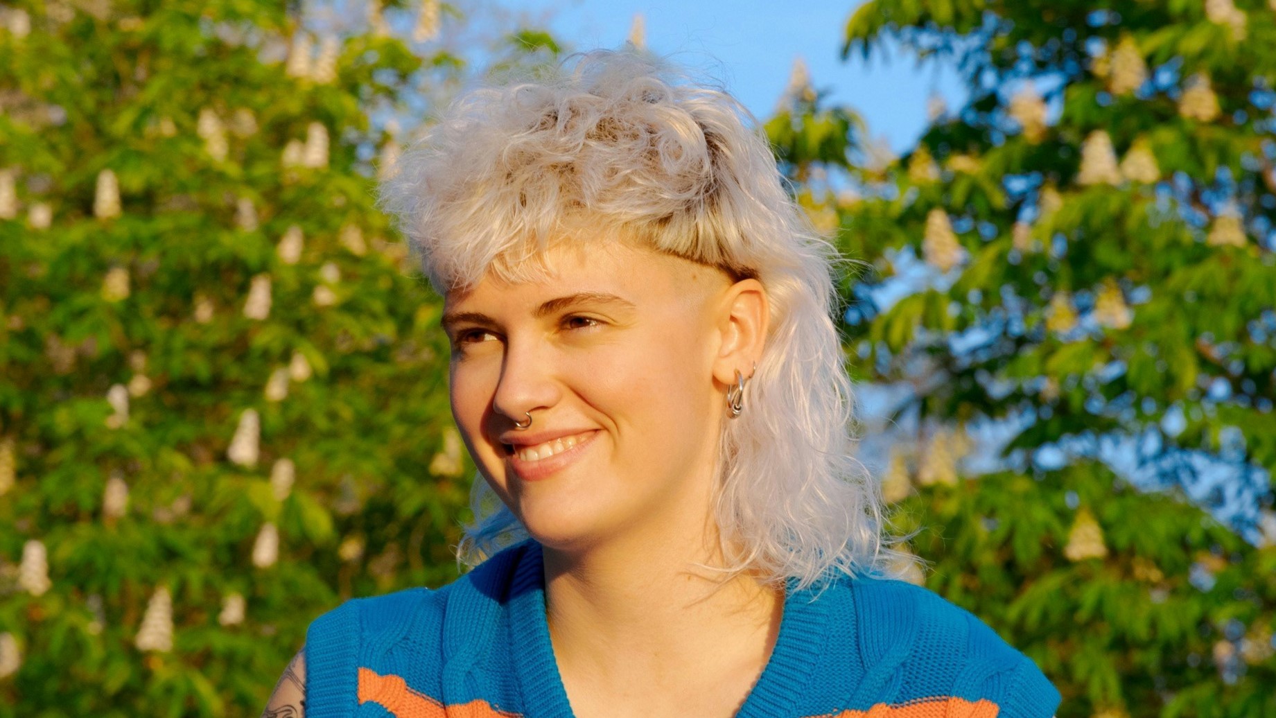 Eine junge Frau mit lockigem, weißblondem Vokuhila-Haarschnitt, die an einem sonnigen Tag draußen sitzt.