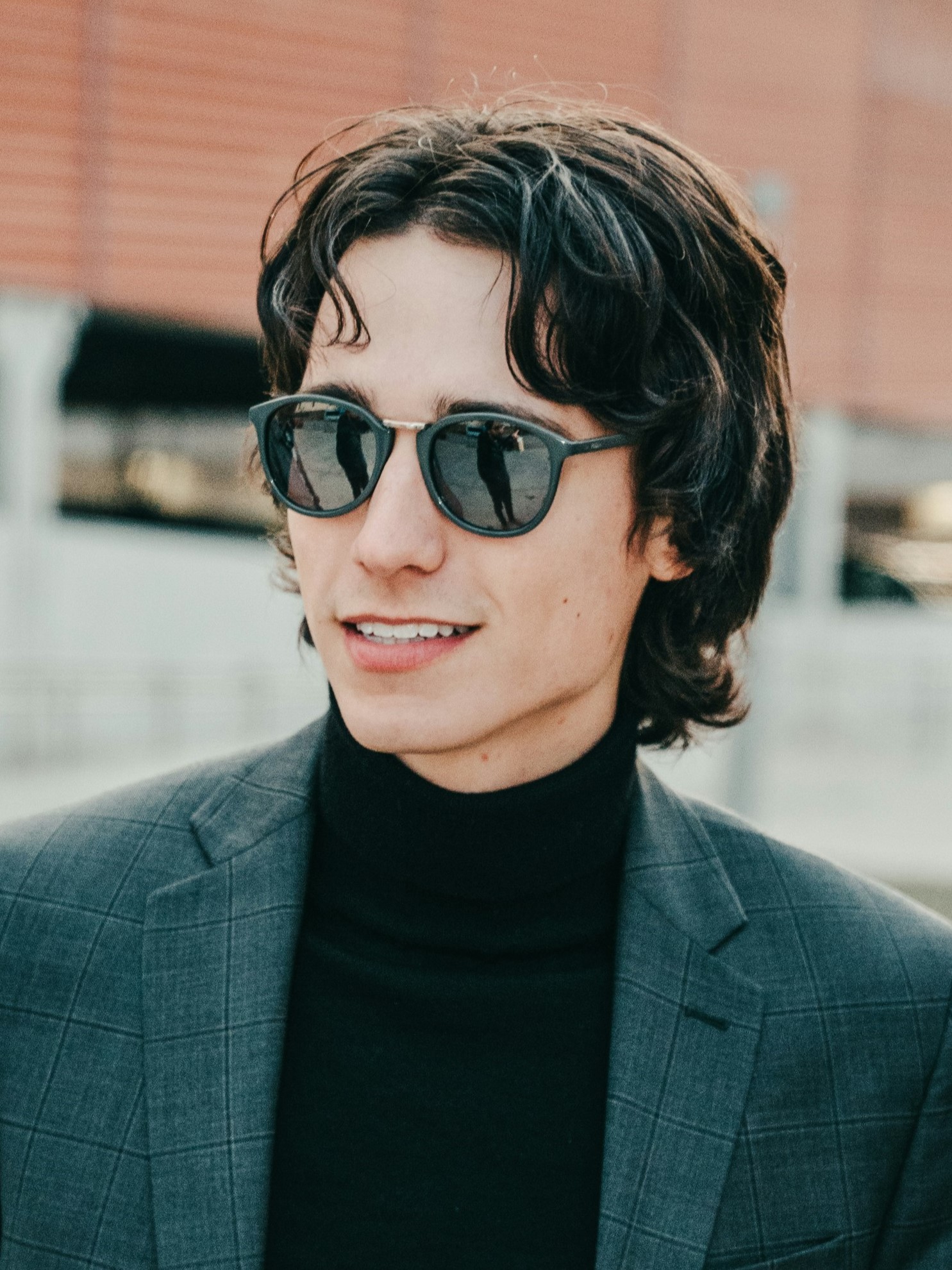 Ein junger Mann mit Retro-Vokuhila, der ein graues Sakko und Sonnenbrille trägt und draußen lächelnd steht.