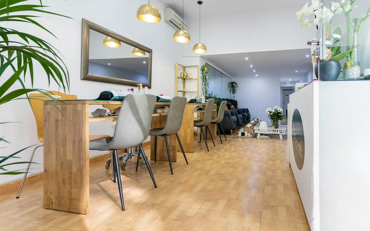 Ein helles Nagelstudio mit langen Holz-Maniküretischen und grauen modernen Stühlen. Goldene Kuppel-Pendelleuchten hängen über den Plätzen, und ein großer Spiegel mit dunklem Rahmen hängt an der weißen Wand.