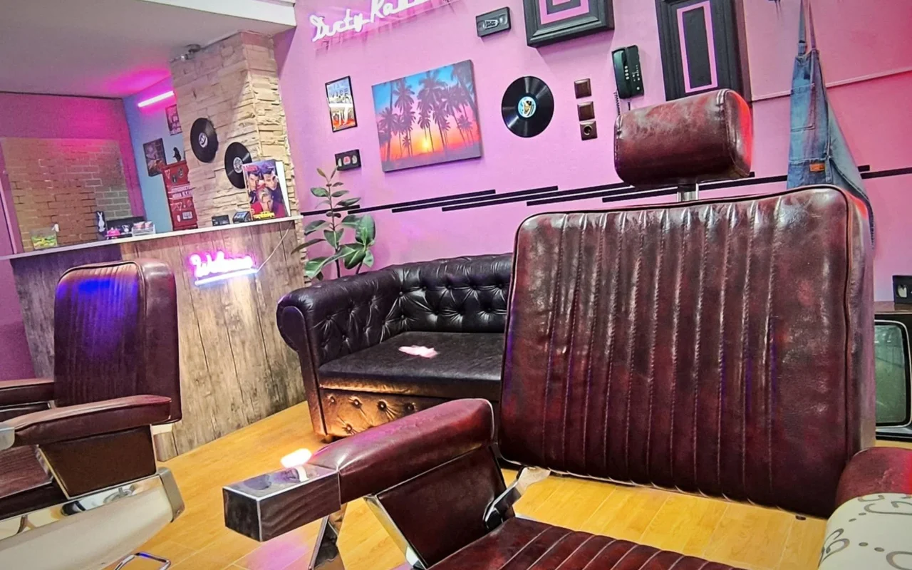 Ein Barbershop im Retro-Stil mit pink-lila Wänden, Neonschriftzügen, Schallplatten als Wanddeko, einem Palmensonnenuntergang-Gemälde, dunklen Leder-Friseurstühlen und einem Chesterfield-Sofa auf warmem Holzboden.