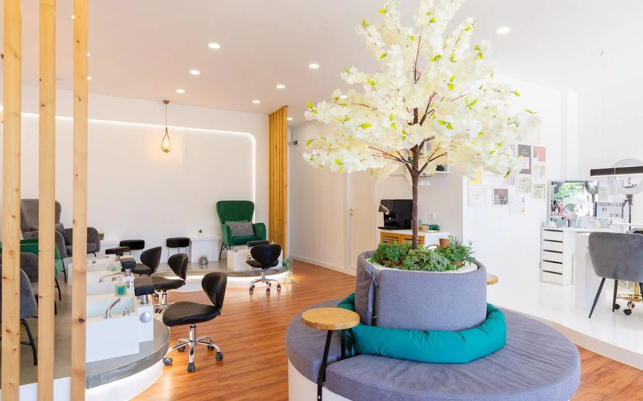 Ein offenes, naturinspiriertes Nagelstudio mit einem großen dekorativen weißen Blütenbaum als Mittelpunkt, smaragdgrünen Samtfauteuils an den Pedikürestationen, Holzlamellentrennwänden und warmem Holzboden.