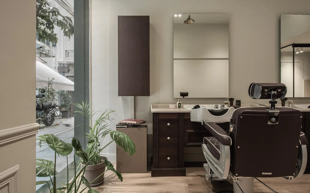 Ein stilvoller Barbershop mit dunkelbraunem Leder-Friseurstuhl, Schrank aus Walnussholz, einem großen Fenster mit Blick auf eine Kolonaki-Straße und einer Monstera-Pflanze, die dem minimalistischen Raum etwas Grün verleiht.