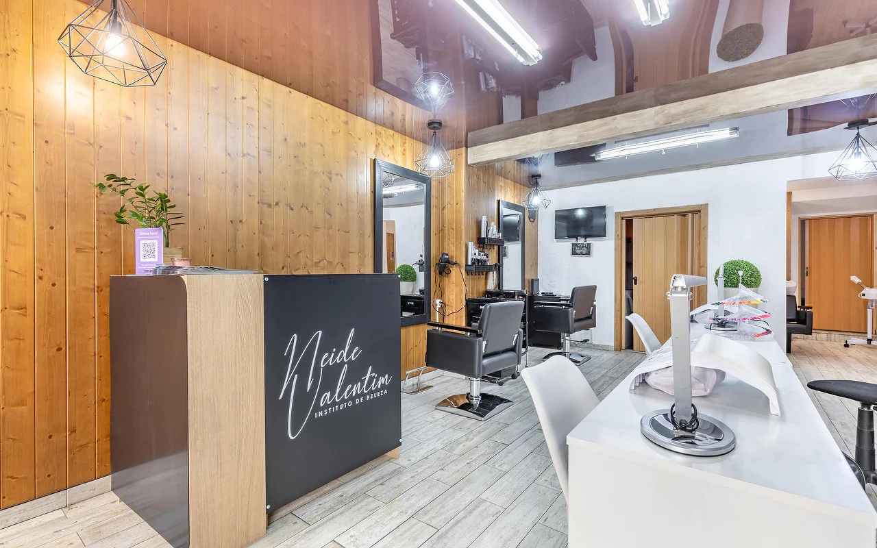 Ein moderner Beauty-Salon mit einer Rezeption links, beschriftet mit „Neide Valentim Instituto de Beleza“. Die Wände sind mit warmem vertikalem Holz verkleidet und großen Spiegeln versehen. Schwarze Frisierstuehle stehen vor den Spiegeln, während auf der rechten Seite weiße Manikürestationen mit weißen Stühlen auf grauem Fliesenboden aufgereiht sind.