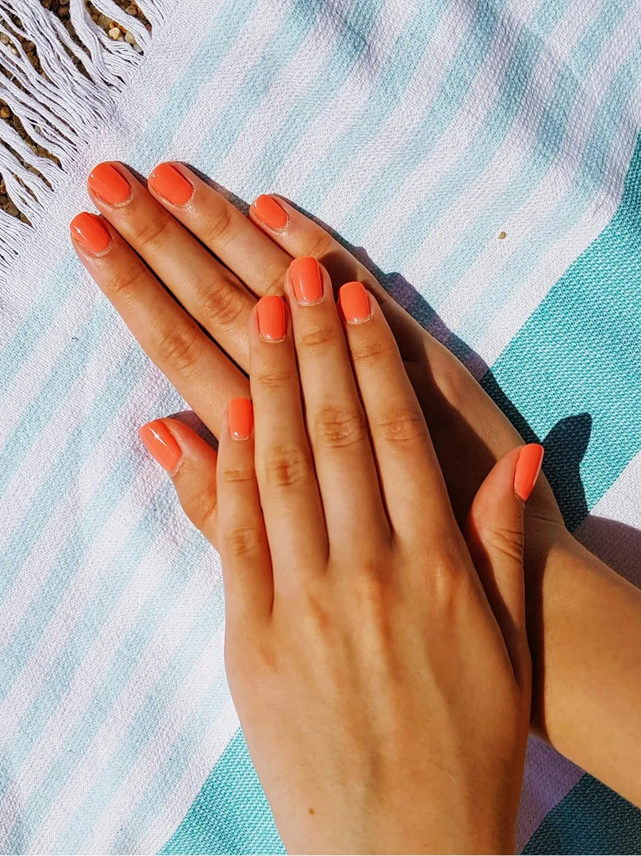 Aufsicht auf zwei Hände mit glänzender oranger Gel-Maniküre auf einem blau-weiß gestreiften Strandtuch