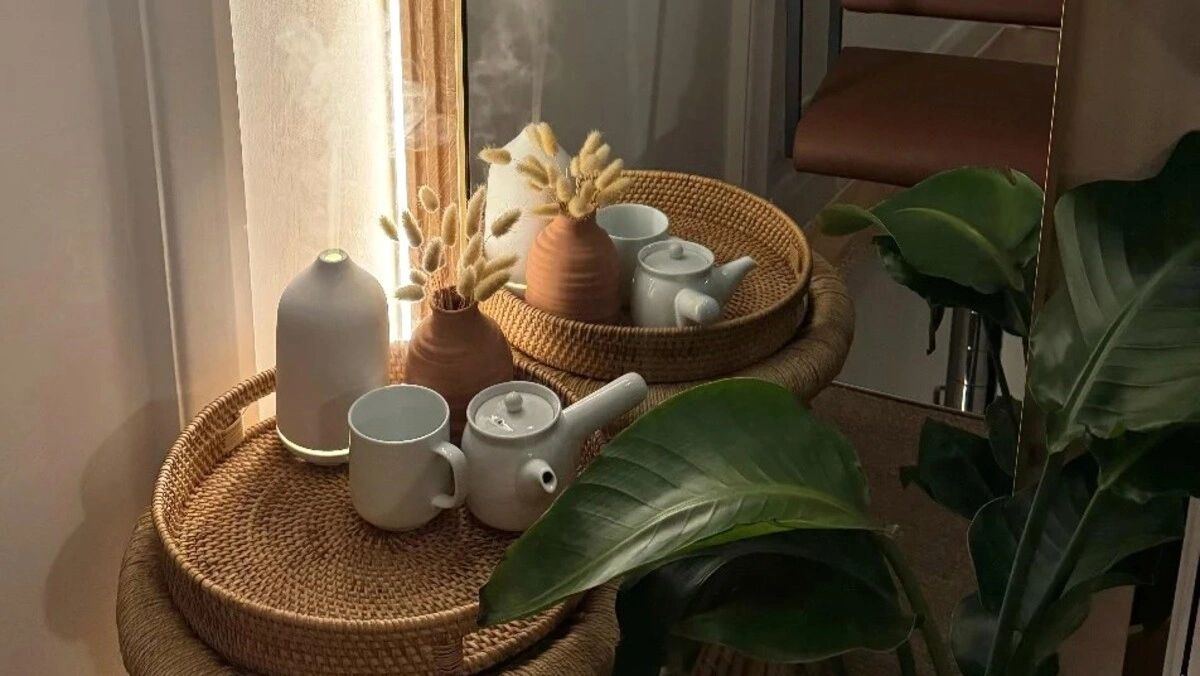 Nahaufnahme eines Korbtabletts mit einem Diffuser und einem Teeservice neben einer großen grünen Pflanze in einem Salon