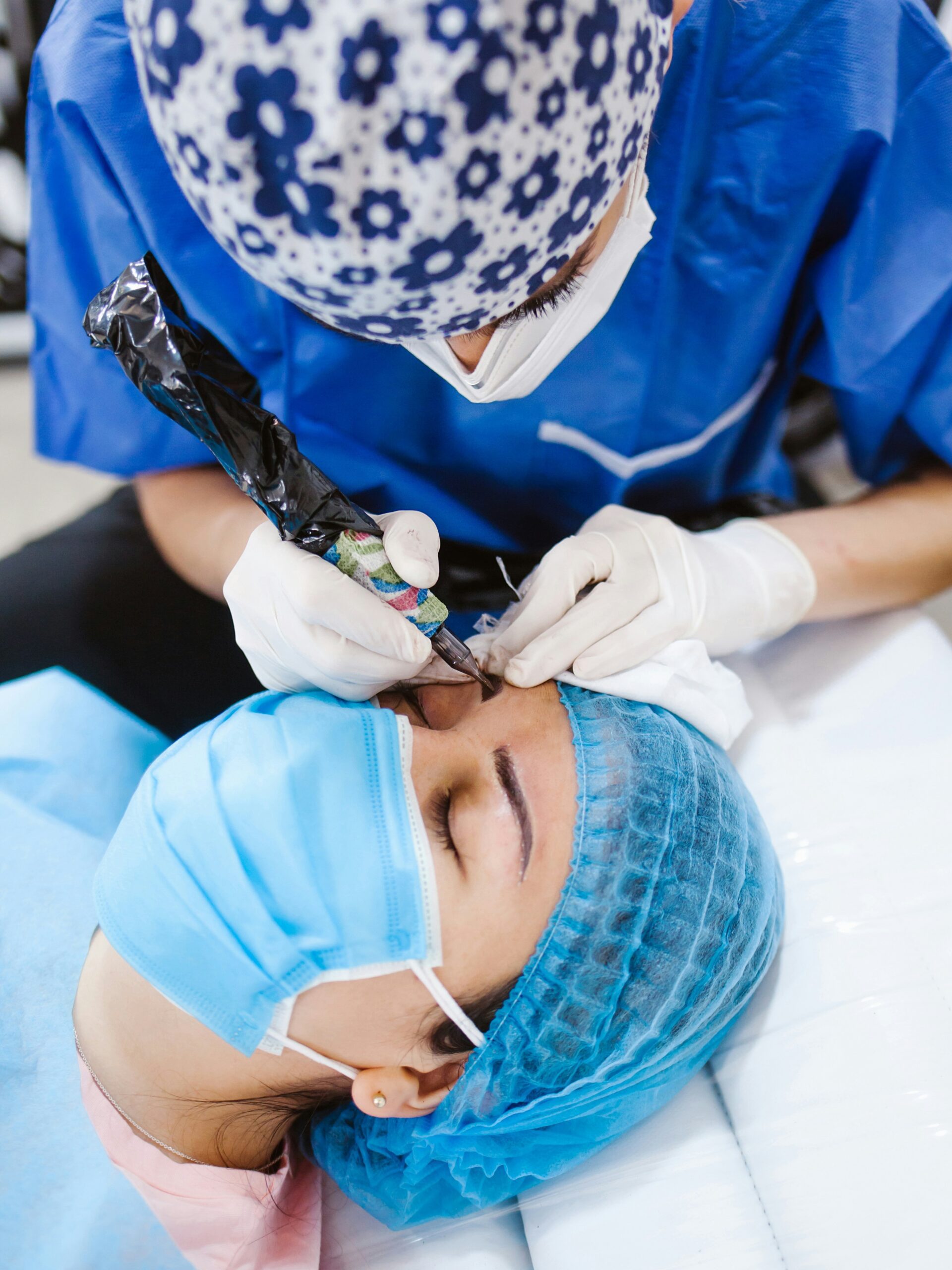 Nahaufnahme einer Frau, die eine Microblading-Behandlung von einer Fachkraft erhält, sie liegt auf der Behandlungsliege und trägt eine Maske