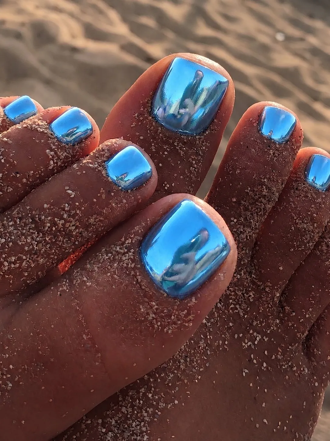 Metallisch glänzende Chrome-Pediküre in Ozeanblau auf sandigen Füßen am Strand