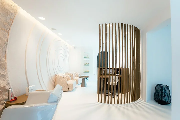 Ein ruhiger, höhlenartiger Spa-Innenraum mit geschwungenen weißen Wänden, eine davon in konzentrischen Wellen ausgearbeitet, sanftes indirektes Licht, cremefarbene Steppsessel und ein geschwungener Holzlamellen-Paravent, der eine ruhige Behandlungsnische abtrennt.