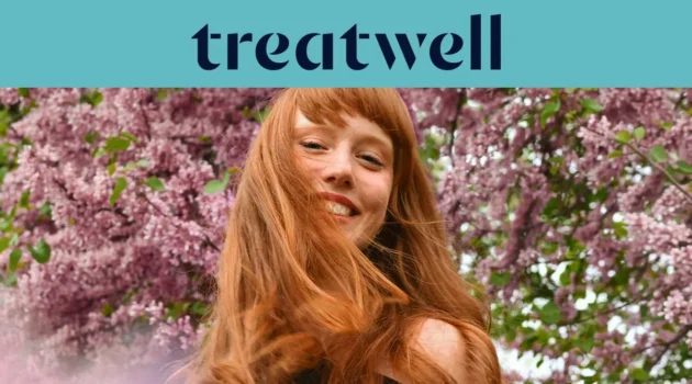 Nahaufnahme einer lächelnden jungen rothaarigen Frau mit Pony, deren Haare vor einem Hintergrund aus rosa Fliederblüten im Wind wehen. Ein hellblaues Banner am oberen Rand zeigt das „Treatwell“-Logo in Dunkelblau.