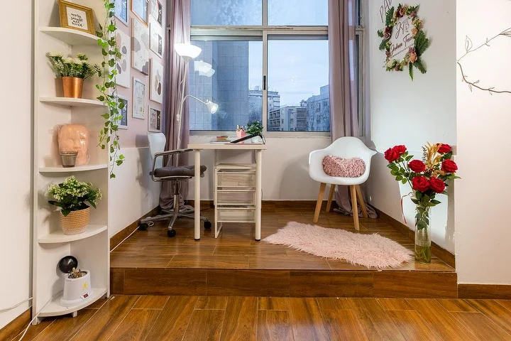 Ein kleines Kosmetikstudio auf einem erhöhten Holzpodest, mit einem weißen Schreibtisch und einer Klemmlampe, einem Bürostuhl und einem Sessel, Eckregalen mit Pflanzen, einer Vase mit roten Rosen und einem Fenster zur Stadt.