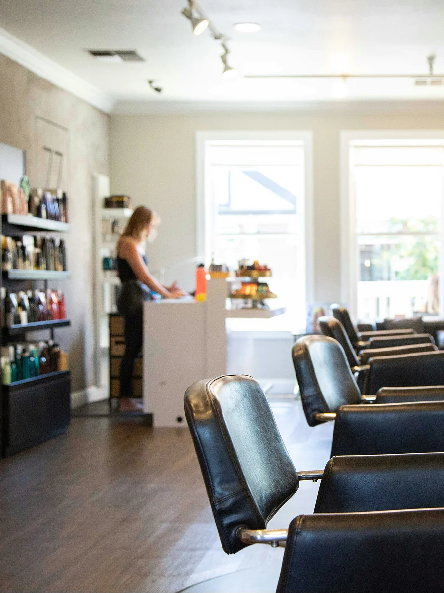 Innenaufnahme eines sauberen, hellen und gut belüfteten Friseursalons mit großen Fenstern, schwarzen Stühlen und Produktregalen