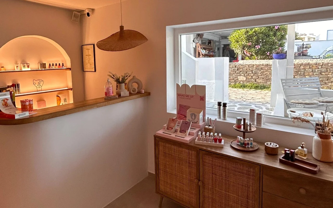 Das Innere von BeautiQ Esthetics and More auf Paros, mit einer sanft beleuchteten gewölbten Nische voller Nagellacke, einer geflochtenen Rattan-Hängeleuchte und einem großen Fenster mit Blick auf eine Steinmauer und einen sonnigen Garten.