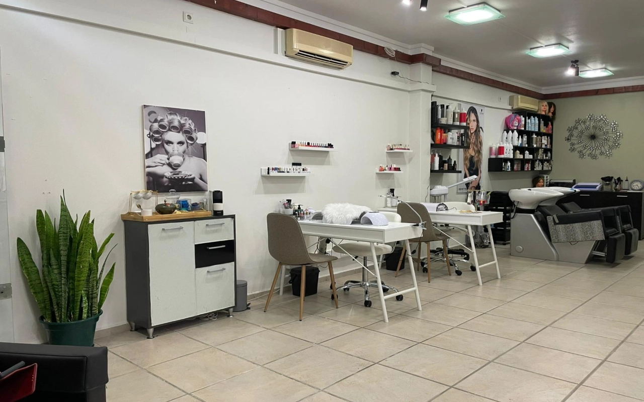 Ein Haar- und Beautysalon mit zwei weißen Maniküretischen und taupefarbenen Stühlen, einem Waschbecken und Produktregalen entlang der Wand, einem hohen Bogenhanf in der Ecke und einer großen Schwarz-Weiß-Fotografie einer Frau mit Lockenwicklern.