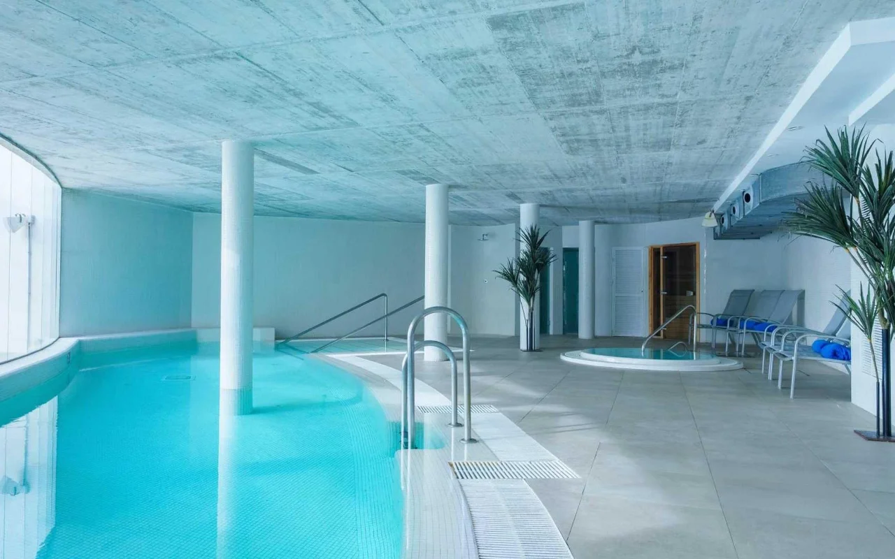 Der Spa-Innenpool von Benalmádena Beach, mit einem geschwungenen türkisfarbenen Becken, einer Sichtbetondecke, einem kleinen Whirlpool und Liegen zwischen hohen Kübelpalmen.