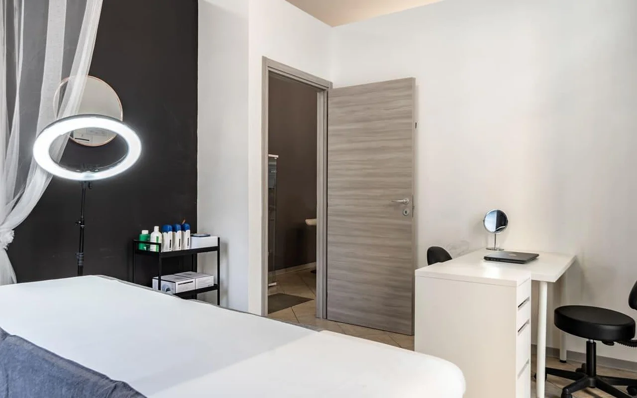 Ein Behandlungsraum bei Coquette Wellness and Beauty, mit einer weißen Liege, einem beleuchteten Ringlicht auf einem Ständer vor einer schwarzen Akzentwand, leichten Vorhängen am Fenster und einem kleinen weißen Schreibtisch mit Schminkspiegel neben einer Tür zu einem eigenen Bad.