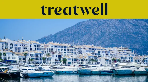 Der Yachthafen Puerto Banús in Marbella, gesäumt von weißen Apartmenthäusern und vertäuten Yachten vor Bergkulisse, unter einem gelben Treatwell-Banner.