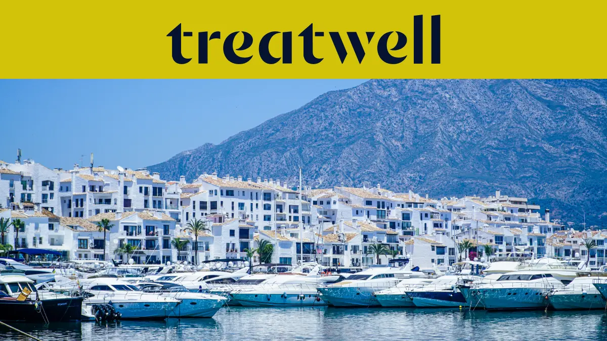 Der Yachthafen Puerto Banús in Marbella, gesäumt von weißen Apartmenthäusern und vertäuten Yachten vor Bergkulisse, unter einem gelben Treatwell-Banner.