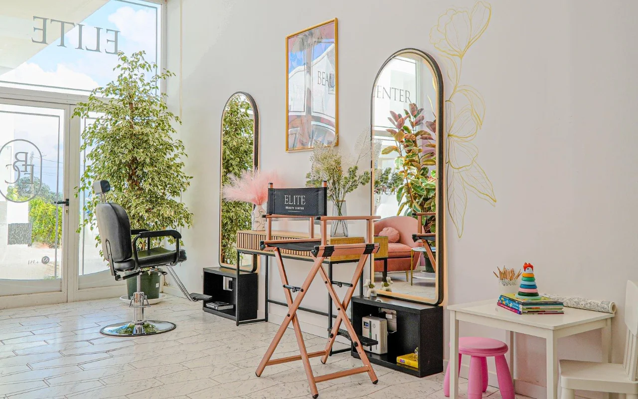Ein heller, luftiger, ganz in Weiß gehaltener Beautysalon mit gewölbten goldgerahmten Spiegeln, einem schwarzen Frisierstuhl und einem klappbaren Regiestuhl, hohen grünen Pflanzen, rosa Akzenten und einer kleinen Kinderecke am Fenster.