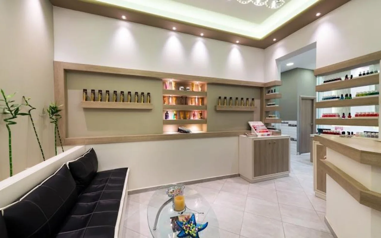 Der cremefarbene Empfang von Fabulous Nails and More in Fira auf Santorini, mit einer schwarzen Lederbank, einem Glastisch, hinterleuchteten Regalen mit Beautyprodukten und sanftem Deckenlicht.
