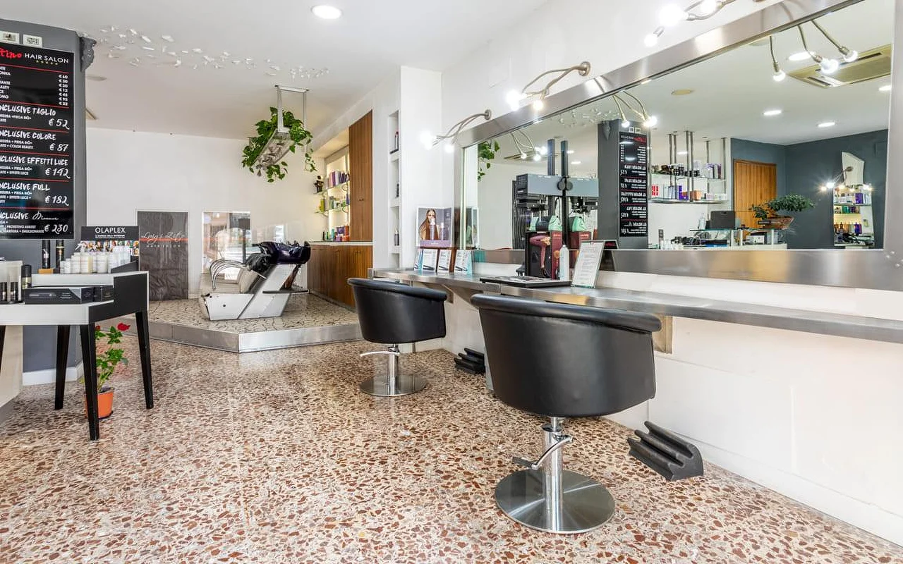 Das Innere des Hair Salon Luigi Zito, mit Terrazzoboden, zwei schwarzen Styling-Stühlen vor einem langen Spiegel, Preistafeln aus Kreide an der Wand, einem Produktdisplay und einem Waschbecken im Hintergrund.
