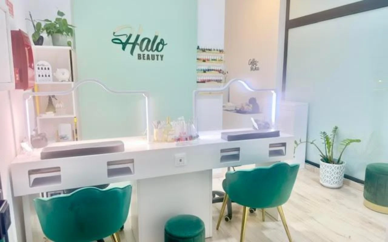 Ein Nagelstudio mit einer mintgrünen Akzentwand mit dem Halo-Beauty-Logo, einem weißen Doppel-Maniküretisch mit beleuchteten Plexiglas-Trennwänden, zwei smaragdgrünen Samt-Tonnensesseln, einem Wandregal voller Nagellacke und Topfpflanzen.
