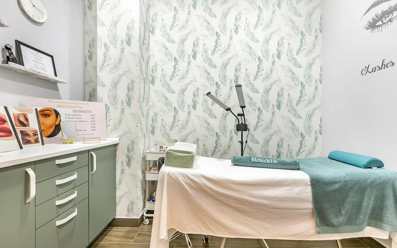 Ein Behandlungsraum bei Masami Belleza Natural in Fuengirola, mit einer Liege, einer Feder-Tapete, einer Lupenlampe und einer Preistafel für Micropigmentierung.