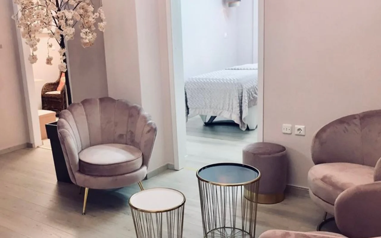 Der zartrosa Wartebereich von Nomikou Antonia Aesthetic Lab auf Syros, mit einem Samtsessel mit gewelltem Rücken, feinen goldenen Beistelltischen, einem blühenden Kirschzweig und einem Behandlungsraum hinter der Tür.