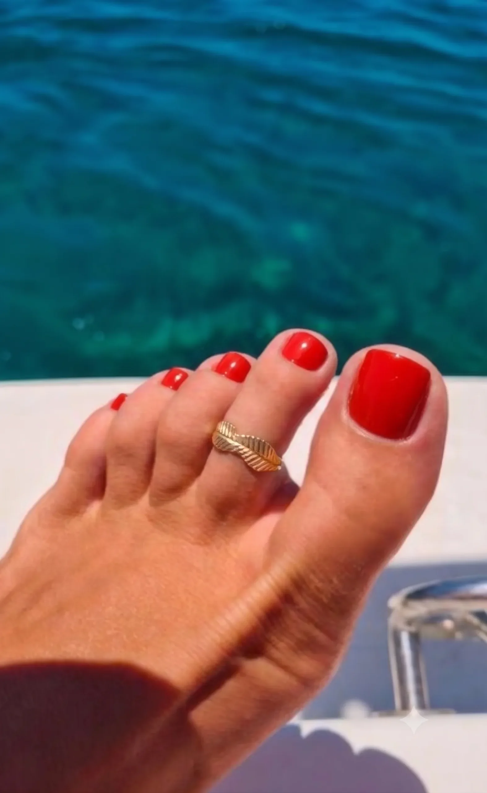 Glänzende tomatenrote Pediküre auf sonnengebräuntem Fuß mit goldenem Zehenring, türkisfarbenes Meer im Hintergrund