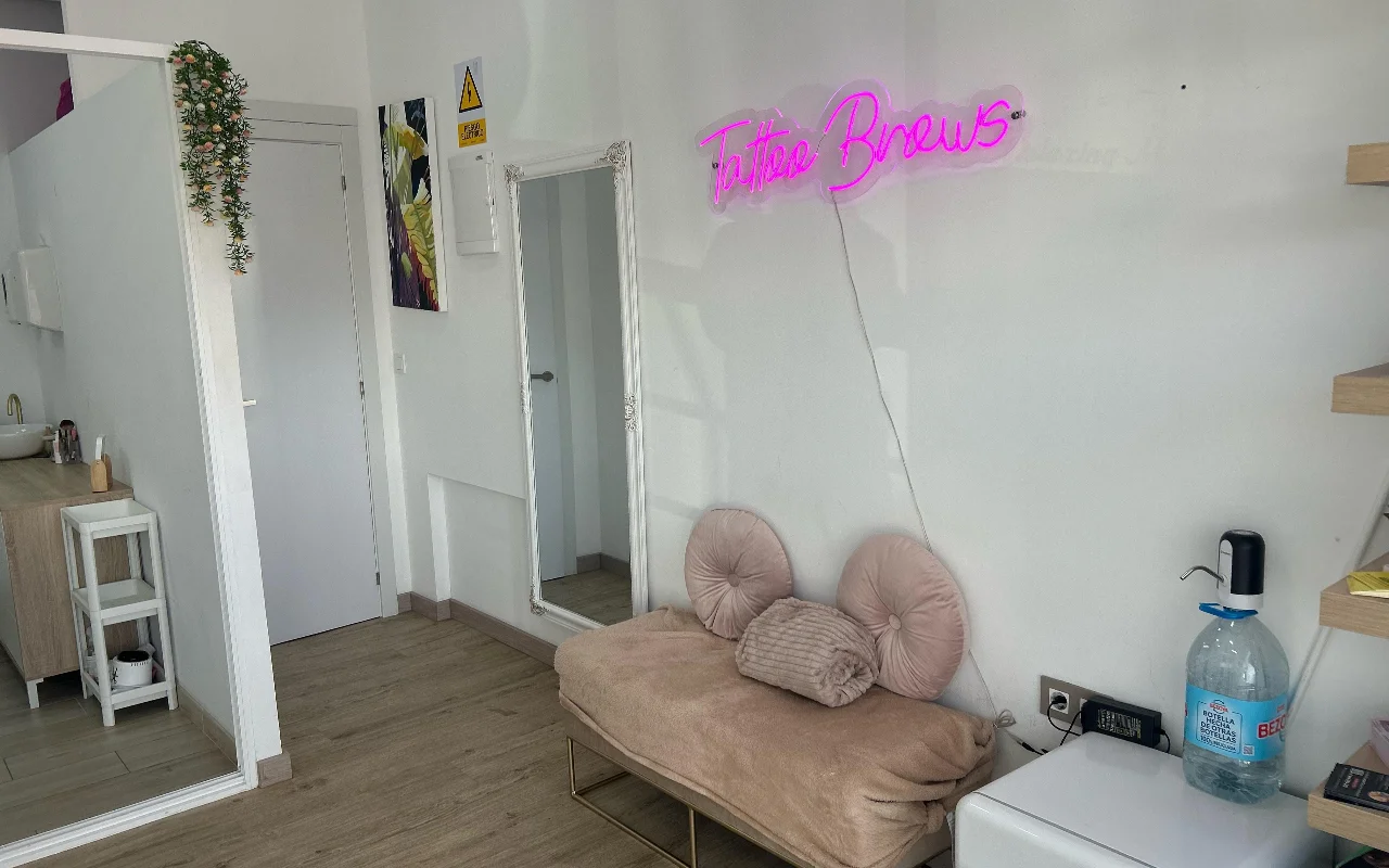 Ein Behandlungsbereich bei YG Beauty Estudio in Torremolinos, mit einer rosa «Tattoo Brows»-Leuchtreklame, einem bodentiefen Spiegel und einer gepolsterten beigen Liege.