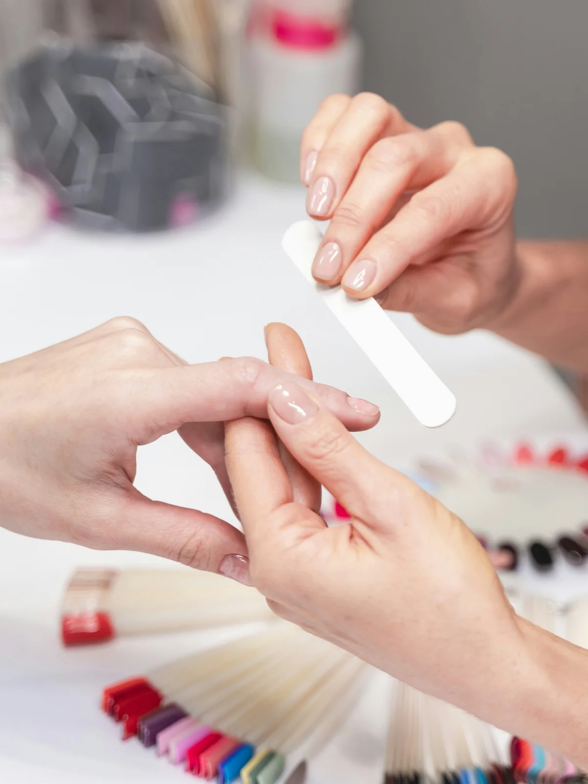 Nahaufnahme einer Nagelstylistin, die die Nägel einer Kundin im Salon sanft feilt, mit Maniküre-Produkten und -Zubehör im Hintergrund