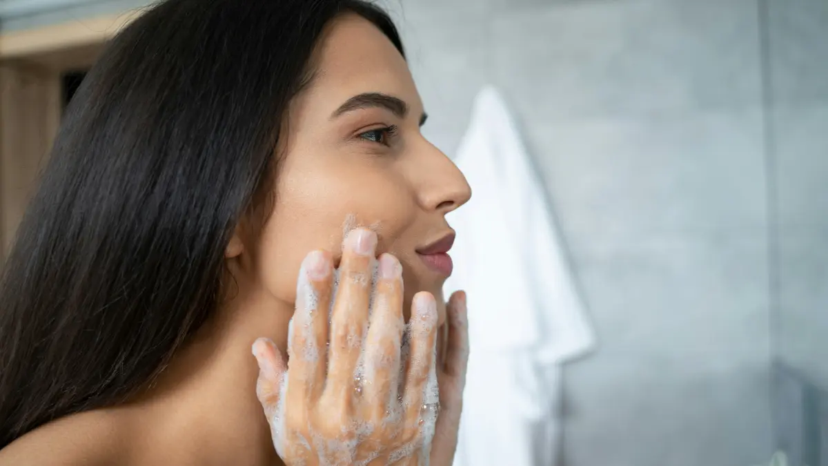 Seitenansicht einer Frau, die sich über dem Waschbecken ein Reinigungsprodukt ins Gesicht einmassiert