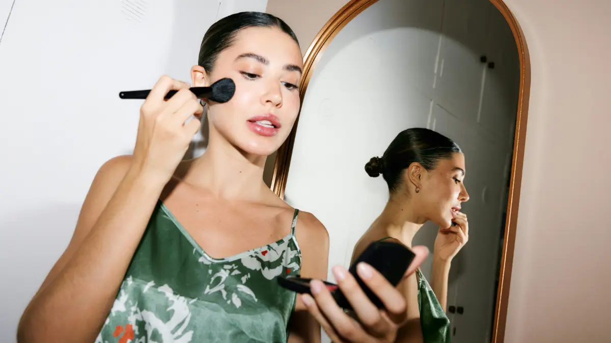Eine glamouröse Frau, fertig zum Ausgehen, schaut in einen Taschenspiegel und trägt mit einem Pinsel Make-up auf