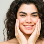 Eine junge Frau mit reiner Haut und dunklen, welligen Haaren lächelt in die Kamera