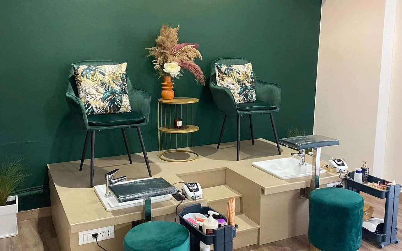 Eine Pediküre-Ecke mit einer tiefgrünen Wand, zwei grünen Samtsesseln mit tropischen Kissen, erhöht auf einer beigen Plattform über Pediküre-Wannen, einem goldenen Beistelltisch mit Pampasgras in einer orangen Vase, und passenden grünen Samthockern.