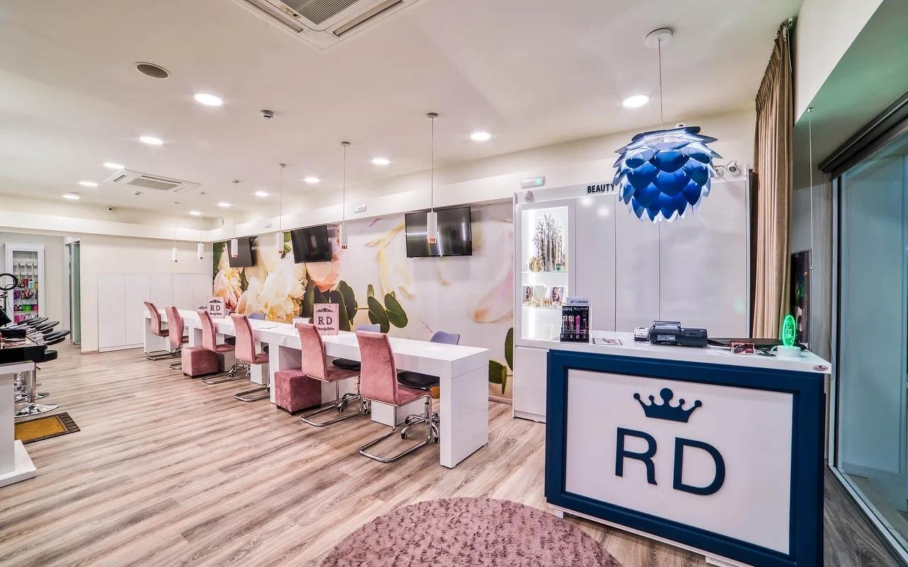 Ein hell-weißer Salon mit einer langen Reihe von Maniküre-Tischen und altrosa Stühlen, einem sanften floralen Wandbild, hellem Holzboden und einer Rezeptionstheke mit einem 'RD'-Kronenlogo.