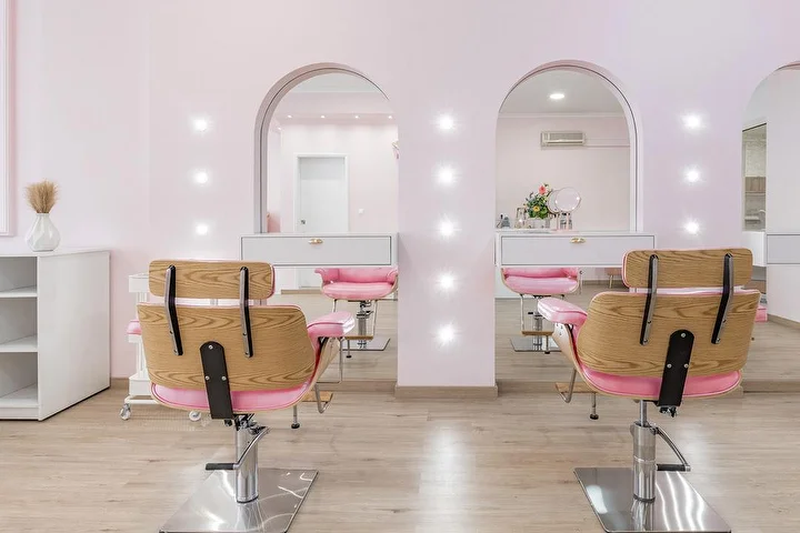 Heller Friseursalon in Zart-Rosa mit Frisierstühlen aus Holz und rosa Stoffbezug vor zwei bogenförmigen, punktbeleuchteten Spiegelnischen, weißen Schränken und Boden in heller Holzoptik.