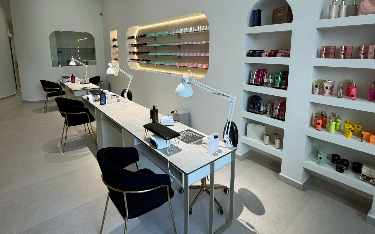 Ein weißes Nagelstudio mit Maniküretischen mit Marmorplatten und weißen Gelenklampen, schwarzen Samtstühlen auf goldenen Beinen, und gewölbten Wandnischen mit Skincare-Produkten und einem hintergrundbeleuchteten Nagellack-Regal.