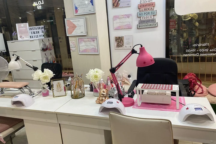 Maniküre-Station eines Nagelsalons mit weißen Tischen, einer kräftig rosa Lampe und LED-Lampen, weißen Rosen und gerahmten Zertifikaten an der Wand, mit dem Namen Color Nails PT auf der Scheibe neben den Öffnungszeiten sichtbar.
