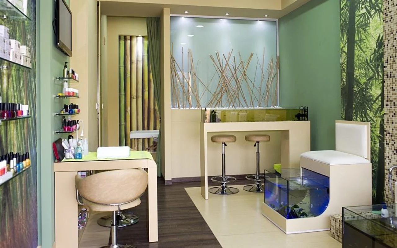 Ein kompakter Salon mit Bambus- und waldgrünem Wanddekor, Wandregalen mit Nagellacken, einem cremefarbenen Maniküretisch und -stuhl, zwei Barhockern und einem blau beleuchteten Glasaquarium für die Fish-Spa-Pediküre.