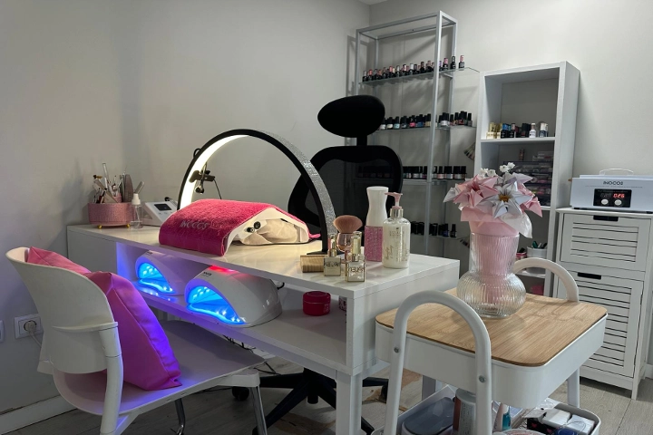 Einzelne Nagelstation mit einem weißen Tisch, einem Ringlicht und zwei blau leuchtenden LED-Lampen, einem schwarzen Stuhl und Regalen voller bunter Lacke, aufgelockert durch eine rosa Vase mit Blumen.