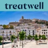 Sonniger Blick über Ibiza-Stadt, mit weiß getünchten Gebäuden, die zur Kathedrale und den alten Festungsmauern von Dalt Vila auf dem Hügel hinaufsteigen. Eine von Bäumen gesäumte Hafenpromenade zieht sich unter einem klaren blauen Himmel durch den Vordergrund.