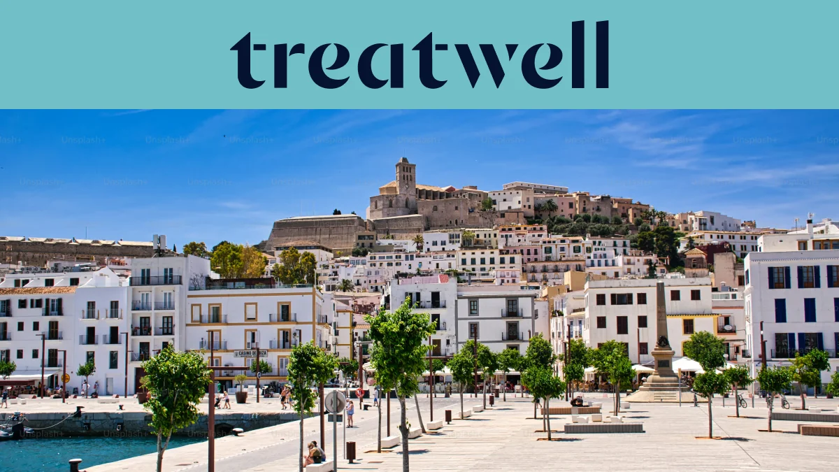Sonniger Blick über Ibiza-Stadt, mit weiß getünchten Gebäuden, die zur Kathedrale und den alten Festungsmauern von Dalt Vila auf dem Hügel hinaufsteigen. Eine von Bäumen gesäumte Hafenpromenade zieht sich unter einem klaren blauen Himmel durch den Vordergrund.