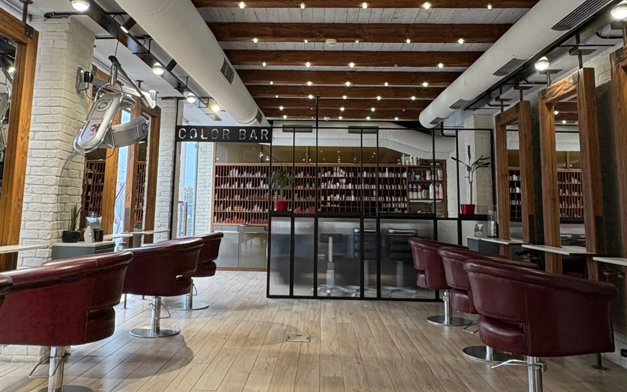 Ein Friseursalon mit freiliegenden Holzdachbalken und weiß getünchtem Ziegelstein, weinroten Lederstyling-Stühlen und einem verglasten Farbraum mit Regalen voller Produkte hinter einem 'Color Bar'-Schild.