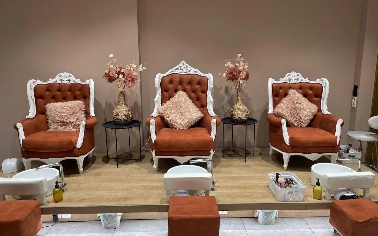 Drei verzierte Sessel mit weißen Rahmen in verbrannt-orangenem Samt, erhöht auf einer Holzplattform über weißen Pediküre-Wannen, mit getrockneten Blumen und weichen rosafarbenen Kissen zwischen den Sitzen.