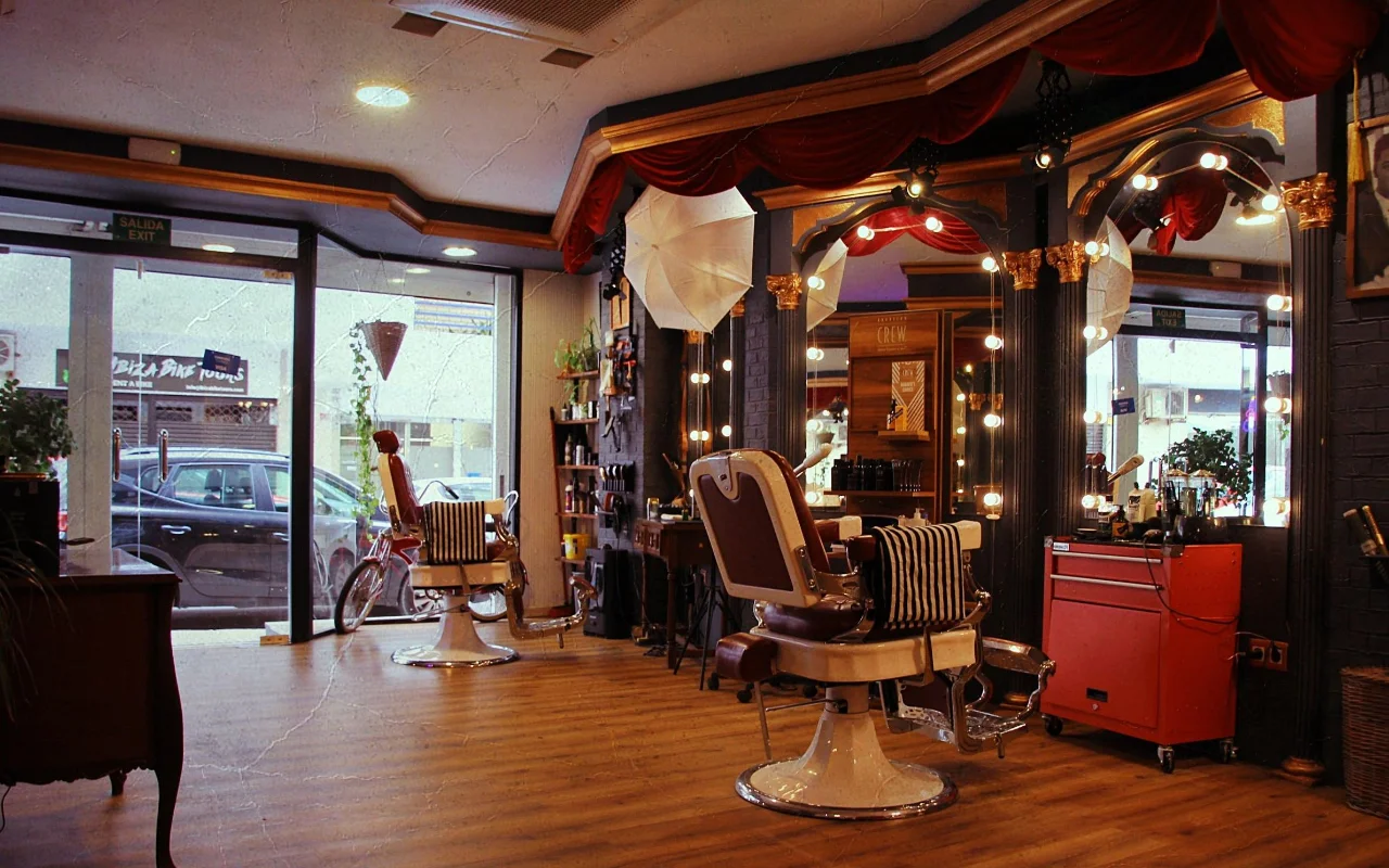 Ein Barbershop im Vintage-Stil mit klassischen Liegesesseln aus Leder auf verchromten Sockeln und gestreiften Handtüchern. Vergoldete Spiegel mit runden Glühlampen und roten Samtvorhängen säumen die Wand, ein roter Werkzeugwagen steht seitlich, und Fenster zur Straße erhellen den Holzboden.