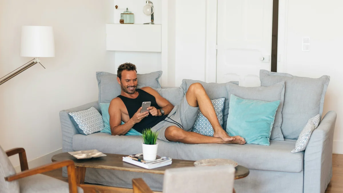 Ein Mann entspannt mit hochgelegten Füßen auf seinem Sofa und lächelt, während er auf sein Handy schaut; er trägt ein schwarzes Trägershirt und eine lockere, weiche graue Shorts